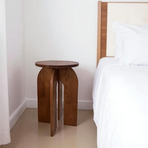 Puede incluir: Una mesa auxiliar de madera con tapa redonda y patas arqueadas. La mesa es de madera marrón claro y está colocada junto a una cama con ropa de cama blanca. El diseño es moderno y minimalista, adecuado para un dormitorio o sala de estar.
