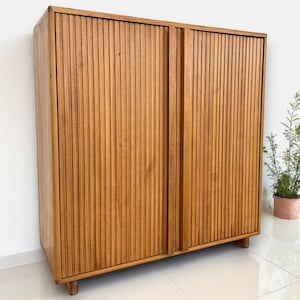 Puede incluir: Un armario de madera con dos puertas correderas. El armario es de madera de color marrón claro y tiene detalles verticales y acanalados en las puertas. El armario es rectangular y se apoya sobre patas cortas de madera. A la derecha del armario se ve una planta en maceta.