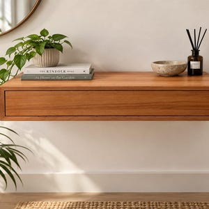 Op de afbeelding: Een rechthoekige, aan de muur gemonteerde zwevende plank van warmgekleurd hout met een geïntegreerde lade. Op de plank staan een groene plant in een pot, twee gestapelde boeken getiteld The Kinfolk Home en At Home in the Garden, een kom en een geurstokjes.