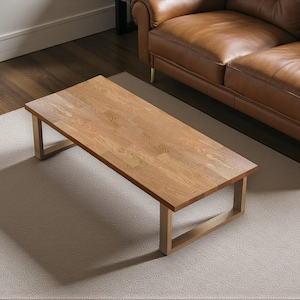 May include: A rectangular wooden coffee table with a light brown finish and a simple, modern design. The table has a light-colored frame and sits on a neutral-toned rug. A brown leather sofa is in the background.