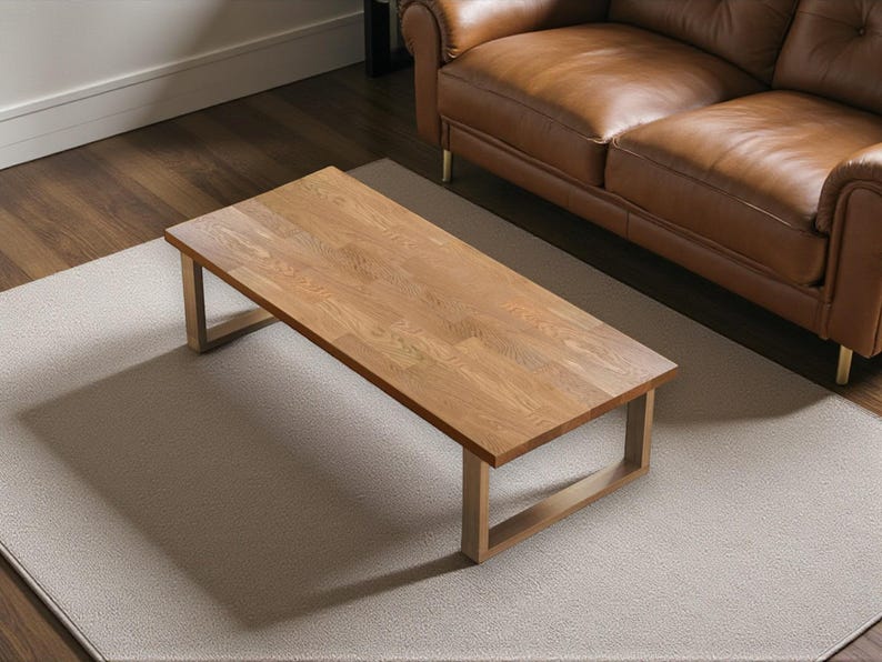 May include: A rectangular wooden coffee table with a light brown finish and a simple, modern design. The table has a light-colored frame and sits on a neutral-toned rug. A brown leather sofa is in the background.