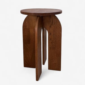 May include: A dark brown wooden side table with a circular top and arched legs. The table has a minimalist design and is made of solid wood. The legs are connected by a central support.