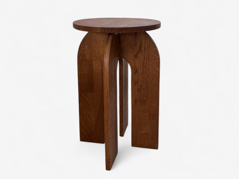 May include: A dark brown wooden side table with a circular top and arched legs. The table has a minimalist design and is made of solid wood. The legs are connected by a central support.