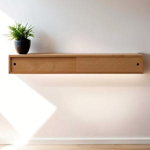 Op de afbeelding: Een licht houten wandplank met twee schuifdeuren. De plank is gemonteerd aan een witte muur en heeft een kleine plant in een zwarte pot erboven.