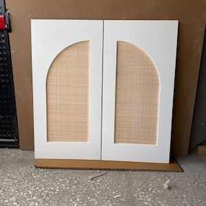 Può includere: Due ante di armadio bianche con pannelli ad arco che incorporano inserti in canna intrecciata. Le ante sono appoggiate su una base di cartone.