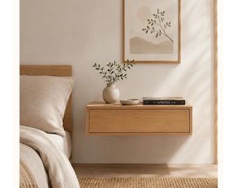 May include: A light oak floating nightstand with a single drawer, mounted on a wall. The top surface holds a small beige ceramic vase with green leafy branches, a shallow beige bowl, and a black book titled The Kinfolk Home.