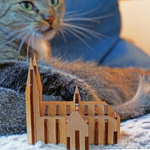 May include: A miniature wooden church model with a light brown color. The model has detailed architectural features, including pointed spires and arched windows. A tabby cat is in the background.
