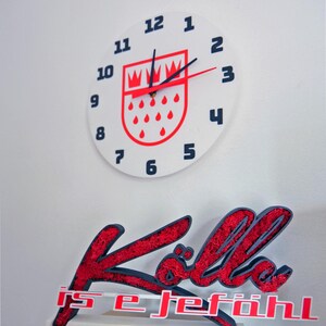 Puede incluir: Un reloj de pared blanco con números negros y manecillas rojas. La esfera del reloj presenta un emblema rojo y blanco. Debajo, un letrero rojo y azul dice "Köln is e jeföhl."