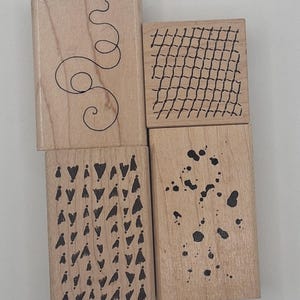 May include: Four rectangular wooden rubber stamps arranged in a square formation. Each stamp features a distinct black ink design on its face: one with a single continuous swirl, one with a grid pattern, one with rows of small triangles, and one with ink splatters.