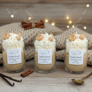May include: Three glass jar candles filled with tan wax, topped with white whipped wax and tan wax spheres. Each label reads Vanilla Bomb, Hand Poured Soy Blend Candle, Net Wt. 7oz/198g. The candles are arranged on a wooden surface.
