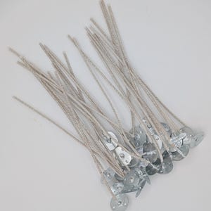 May include: A collection of candle wicks with metal tabs. The wicks are made of a light beige braided material, and the metal tabs are silver-colored. The wicks are arranged in a scattered pattern against a white background.