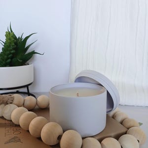May include: A gray candle in a gray tin with a lid. The candle is surrounded by wooden beads.