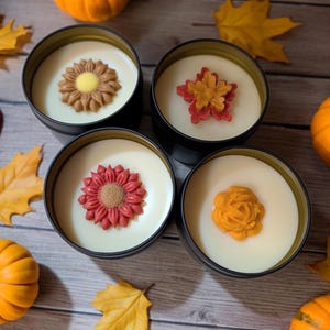 Puede incluir: Cuatro velas de lata negras con diseños florales. Cada vela tiene un diseño floral diferente en tonos marrones, rojos y naranjas. Las velas están rodeadas de calabazas y hojas de otoño sobre una superficie de madera.