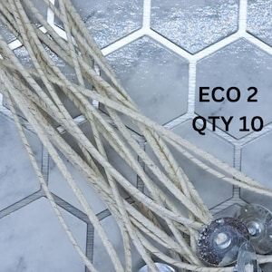 May include: A close-up of ten white cotton candle wicks with metal bases. The wicks are arranged in a pile on a gray and white tiled surface. The text "ECO 2 QTY 10" is visible in the image.