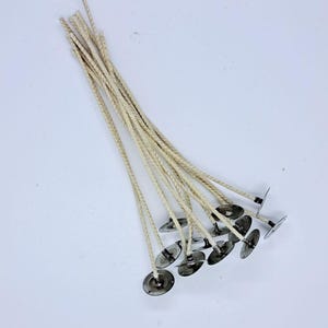May include: A collection of ten off-white braided cotton candle wicks, each attached to a circular silver-colored metal sustainer base. The wicks are arranged in a fan-like cluster, with the metal bases grouped together at the bottom.