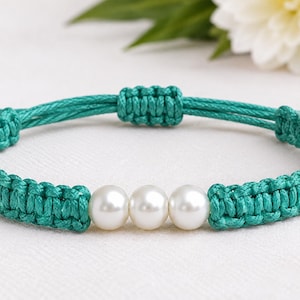 May include: A teal cord bracelet featuring a macrame knot pattern and three smooth white spherical pearls centered in the design. The bracelet includes an adjustable sliding knot closure with two knotted ends on each side of the cord band.