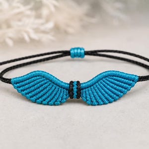 May include: A macrame bracelet featuring a central wing-shaped design crafted from bright turquoise cord. The wings are accented with a small black cord detail in the center. The bracelet is held together by a thin black cord with adjustable knots.