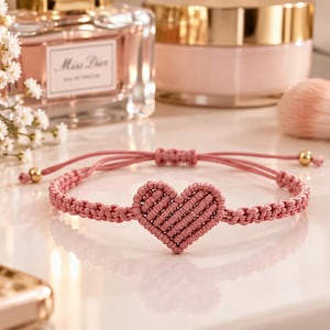 Könnte beinhalten: Ein altrosa Makramee-Armband mit einem zentralen herzförmigen Knotenmuster. Das Armband besteht aus einem dünnen Kordelband mit einem verstellbaren Schiebeknotenverschluss und zwei kleinen goldfarbenen Metallperlen an den Enden.
