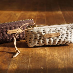 May include: Two rectangular zipped pouches, one dark brown and one off-white, featuring a quilted texture with visible running stitches. Each pouch has a brass-toned zip, a leather pull cord, and a small rectangular tag labelled Chandra's House.
