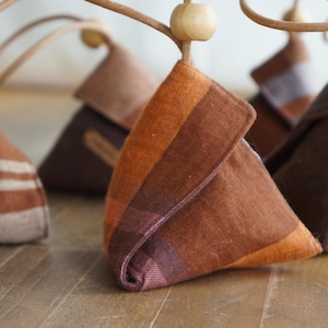 May include: Close-up of several triangular fabric ornaments in shades of brown, orange, and purple. Each ornament has a wooden bead and a leather loop for hanging. The fabric has a plaid pattern.
