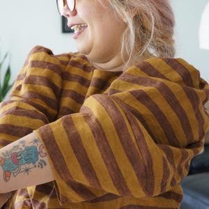 Könnte beinhalten: Ein senfgelbes und braun gestreiftes Oberteil mit Puffärmeln. Das Oberteil wird mit einem senfgelben Cordrock kombiniert. Ein farbenfrohes Tattoo ist auf dem Arm zu sehen.