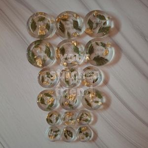 May include: Clear resin buttons with embedded green leaves and gold flakes. The buttons are round and have a flat back with a loop for sewing. The text "your stitcher friend" is visible in the image.