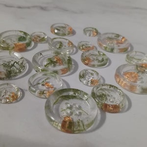 May include: A collection of 14 clear resin buttons with gold flecks and green botanical inclusions. The buttons are various sizes and shapes.