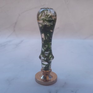 May include: A decorative wax seal stamp with a clear handle filled with green moss. The stamp has the words "Your Stitcher Friend" etched into the handle. The base of the stamp is gold-colored.