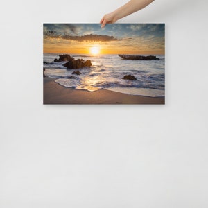 May include: A framed canvas print of a beach scene at sunset. The sun is setting over the ocean, casting a golden glow on the water and sand. Dark rocks are in the foreground, and the sky is filled with clouds.
