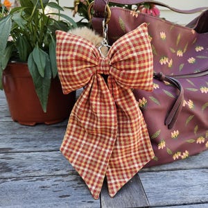 May include: A large fabric bow with a red, yellow, and white gingham check pattern, attached to a silver-toned metal lobster clasp. The bow features two long, pointed tails and is suspended from a dark maroon handbag with a floral print pattern.
