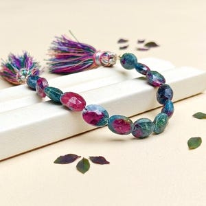 May include: A beaded bracelet featuring faceted oval-shaped ruby and zoisite gemstones in shades of deep magenta and forest green. The bracelet is finished with two decorative multicolored fiber tassels attached to silver-toned metal caps.