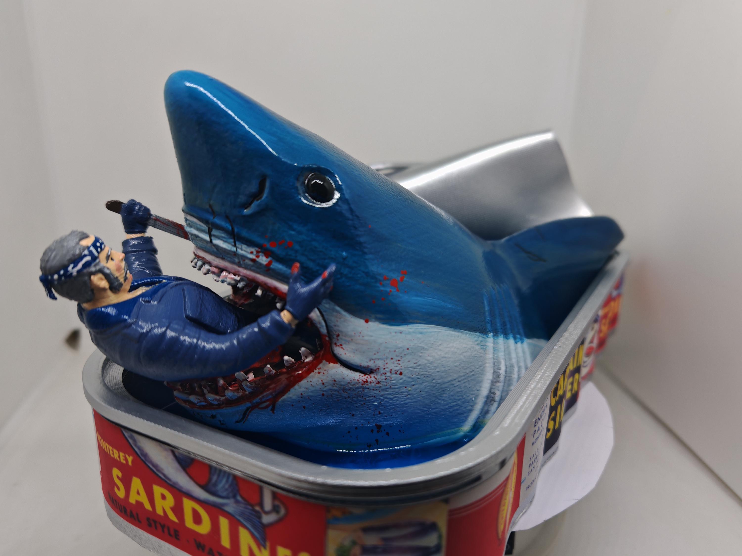 Op de afbeelding: Een handbeschilderd beeldhouwwerk met een grote blauwe haai die uit een metalen sardineblikje komt. Een figuur in een blauw uniform bevindt zich in de open bek van de haai en houdt een klein gereedschap vast. Het blikje heeft een rood etiket.