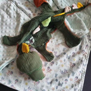 Könnte beinhalten: Ein grünes und orangefarbenes Plüschdrachenspielzeug mit gelben und beigefarbenen Akzenten. Der Drache liegt auf einer hellblauen und weiß gemusterten Decke mit einem kleinen weißen Etikett, auf dem "M.E.S." steht.