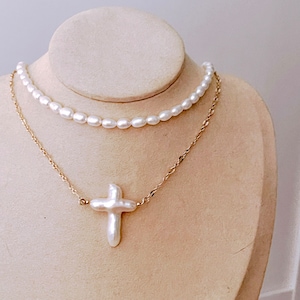 以下が含まれることがあります： マネキンに展示された2連ネックレスセット。 上段は繊細な白い真珠の連なりです。 下段は、大きな白い真珠の十字架ペンダントが付いたゴールドチェーンが特徴です。