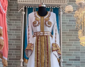 May include: A white maxi dress with gold and burgundy embroidery. The dress has a long, flowing skirt and long sleeves. The embroidery is intricate and detailed, and the dress is perfect for a special occasion.