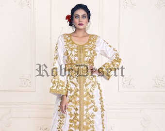 Op de afbeelding: Een witte kaftan met gouden borduurwerk en pailletten. De kaftan heeft lange mouwen en een riem. Het model draagt een bloem in haar haar.