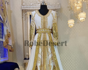 May include: A white and gold kaftan with intricate embroidery and sequins. The kaftan has a long, flowing skirt and a fitted bodice. The fabric is sheer and delicate, and the overall effect is elegant and luxurious.