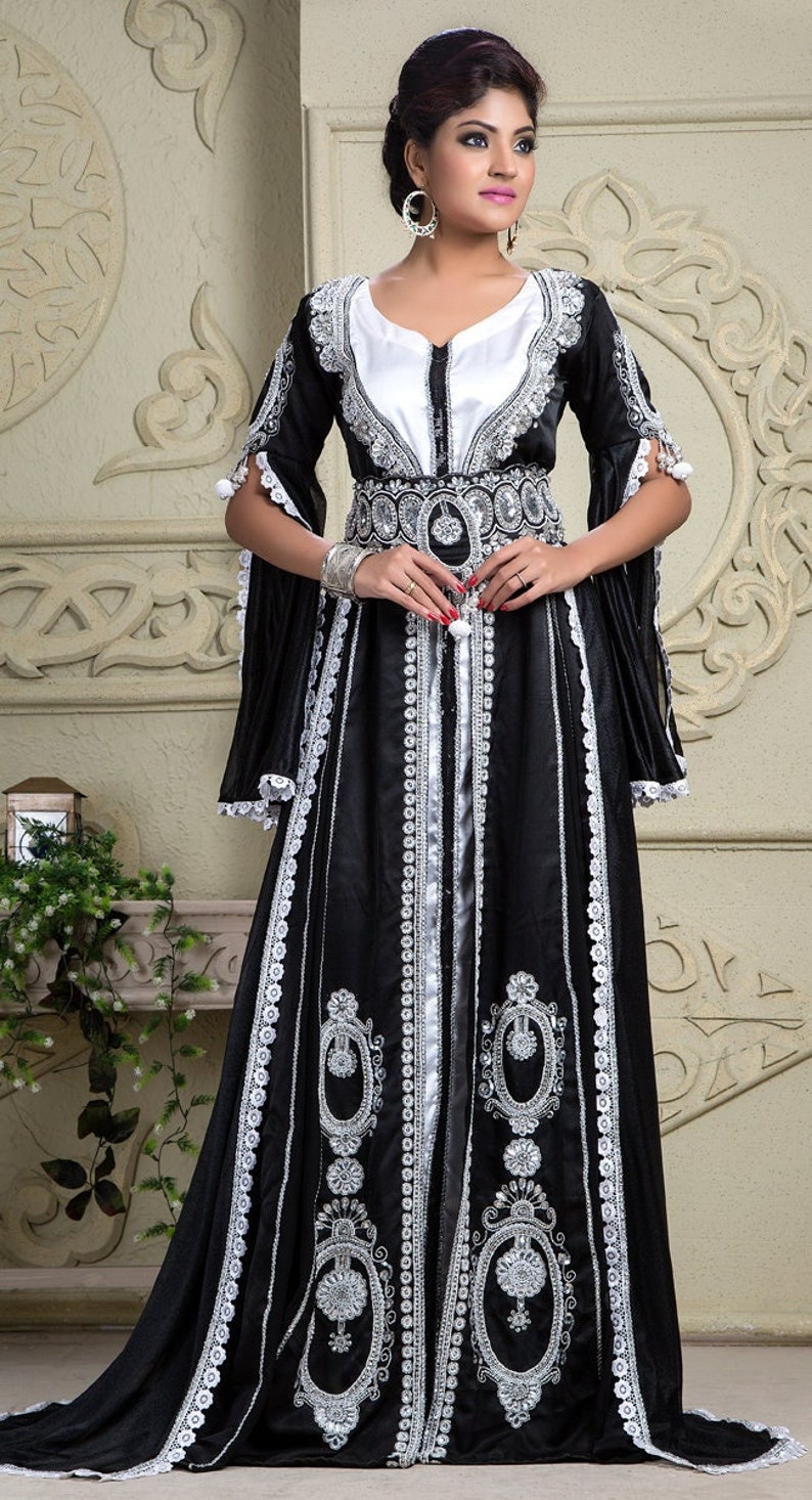 Op de afbeelding: Een zwarte en zilveren kaftan met lange mouwen en ingewikkelde borduursels. De kaftan heeft een witte voering en een zilveren riem.