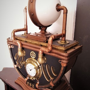 Könnte beinhalten: Eine Steampunk-Uhr mit einer Kupfer- und Messing-Oberfläche. Die Uhr hat ein rundes Zifferblatt mit römischen Ziffern und eine große, weiße, kugelförmige Leuchte oben. Die Uhr ist auf einem Holzsockel mit der Nummer "D52" auf der Vorderseite montiert.