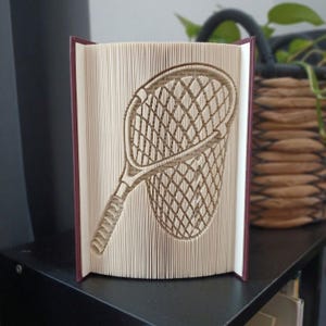 Peut inclure: Une sculpture de livre avec des pages pliées formant la silhouette d'une raquette de tennis et d'une balle de tennis. Le livre possède une couverture bordeaux foncé et les pages sont de couleur crème, créant une représentation en relief.