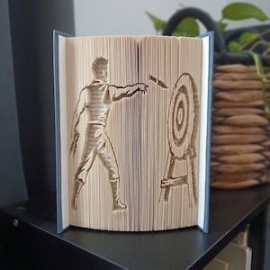 Pode incluir: Uma escultura de livro dobrado apresentando a silhueta de uma pessoa lançando uma faca em um alvo circular. As páginas são dobradas com precisão para criar a imagem, com as capas do livro visíveis nos lados esquerdo e direito da peça.