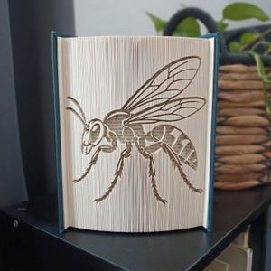 May include: A folded book art sculpture featuring a detailed wasp silhouette created by precisely folding the pages of a book. The insect design displays clear wings, segmented body parts, and thin legs formed by the edges of the paper pages.