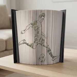 Könnte beinhalten: Dekorative Buchskulptur mit einem Fußballspieler, der einen Ball tritt. Das Bild entsteht durch das Falten der Buchseiten, wobei die Figur des Spielers in Grün- und Grautönen gehalten ist. Das Buch steht auf einer hellen Holzoberfläche.
