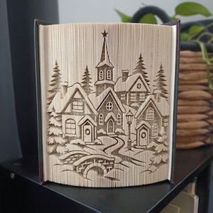 Op de afbeelding: Een gevouwen boekkunstsculptuur met een gedetailleerd winterdorp. De pagina's zijn nauwkeurig gevouwen om een centrale kerk met een toren, omliggende huizen met puntdaken, groenblijvende bomen en een kleine boogbrug te onthullen.