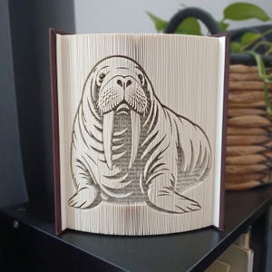 May include: A book sculpture featuring a walrus design created by folding the pages of a book. The walrus is depicted with prominent tusks, whiskers, and flippers, with the text of the book pages visible through the folded paper structure.