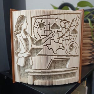 Peut inclure: Une sculpture en livre plié représentant la silhouette d'une personne debout à côté d'une carte météorologique. La carte affiche des symboles pour un soleil, des nuages, de la pluie et un ouragan. Les pages sont pliées pour former ces motifs.