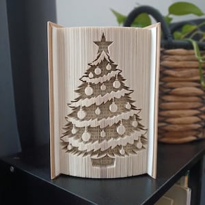 Op de afbeelding: Een gevouwen boekkunstsculptuur met een kerstboomontwerp, gemaakt door de pagina's van een boek nauwkeurig te vouwen. De boom is versierd met ronde ornamenten en bekroond met een ster, waarbij de tekst op de papierranden zichtbaar is.