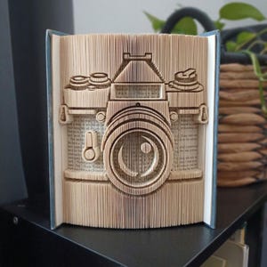 Può includere: Una scultura di libro con pagine piegate che formano la sagoma di una fotocamera vintage. I bordi della carta sono piegati con precisione per creare il corpo della fotocamera, un obiettivo centrale e ghiere superiori, rivelando testo stampato.
