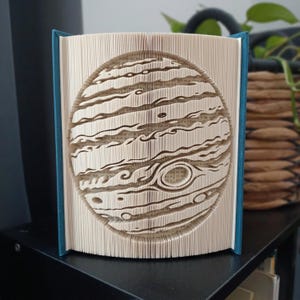 Peut inclure: Une sculpture en livre plié représentant un relief circulaire de la planète Jupiter. Les pages sont précisément pliées pour former les bandes caractéristiques de la géante gazeuse et la Grande Tache rouge. Le livre a un dos bleu sarcelle.