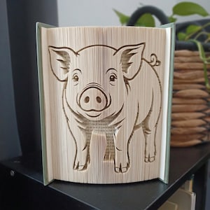 Peut inclure: Une sculpture de livre représentant un cochon créée en pliant les pages d'un livre à couverture rigide. Le cochon est représenté debout avec son museau, ses oreilles et sa queue en tire-bouchon clairement définis par les plis du papier.